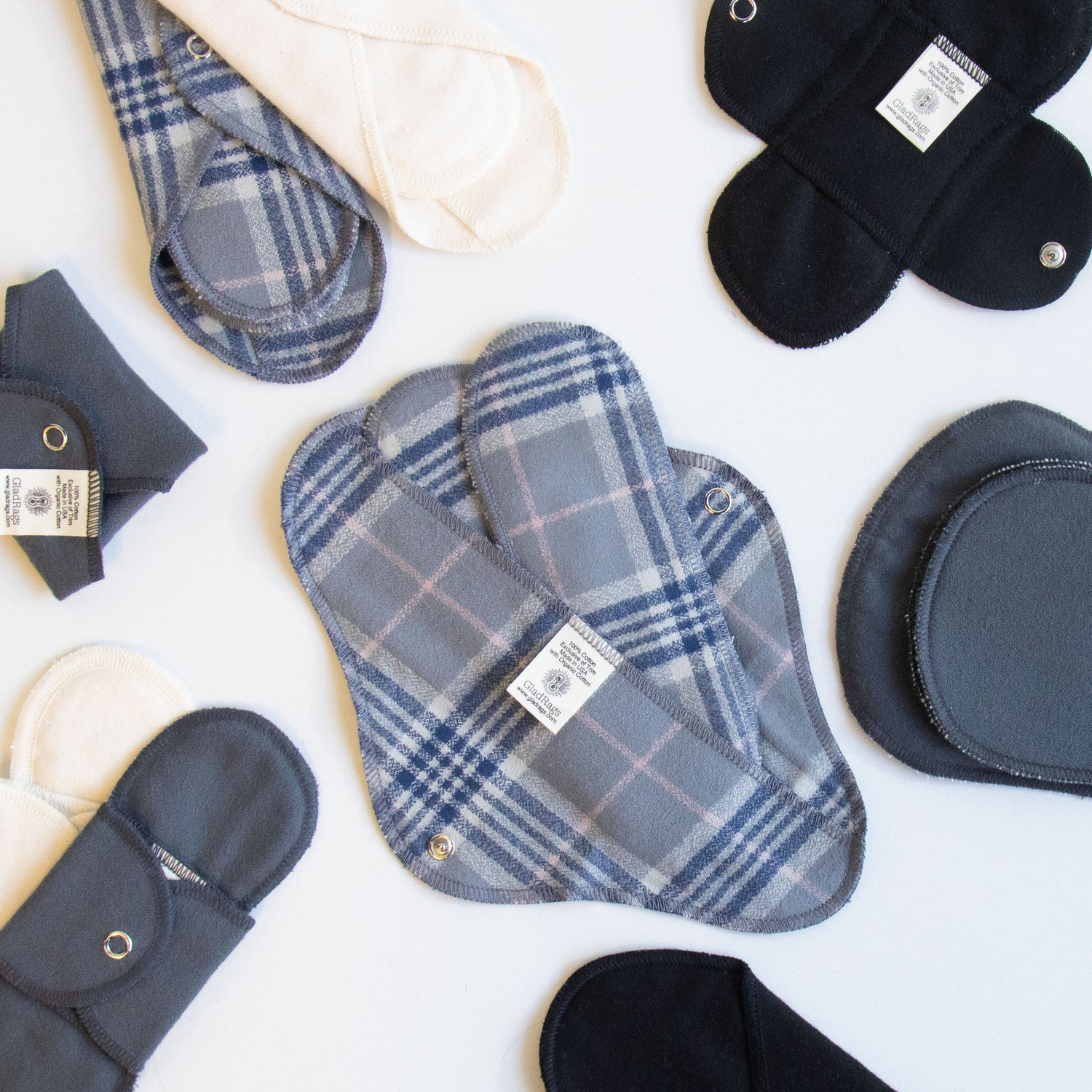 cream, grey plaid, grey, and black organic reusable pads of various sizes on a white background