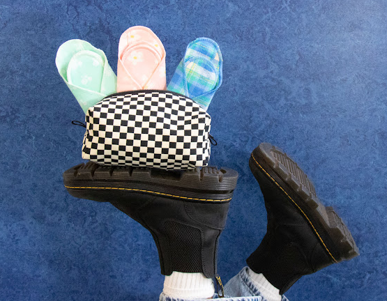 upside-down ankles and feet in black boots. Placed on the sole of one boot is a black-and-white checked GladRags Weekender Bag with three cloth pads sticking out: a mint green one, a light pink one, and a blue plaid one.