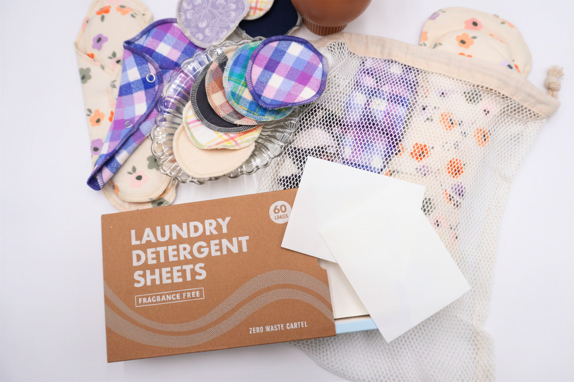 Flat lay of reusable menstrual care products and laundry supplies, including cloth pads, reusable cotton rounds, a mesh wash bag, and fragrance-free laundry detergent sheets. Various floral and plaid fabric patterns are displayed on a white background.